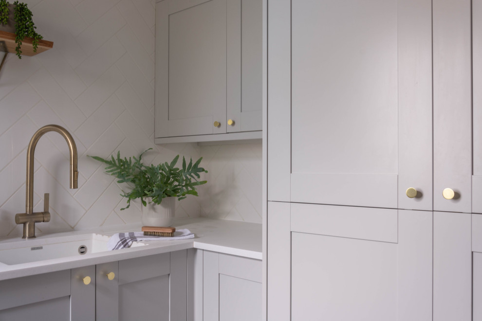 Inspiration for a small modern l-shaped utility room in Cambridgeshire with an undermount sink, shaker cabinets, grey cabinets, solid surface benchtops, white splashback, ceramic splashback, grey walls, porcelain floors, a stacked washer and dryer, grey floor and white benchtop.