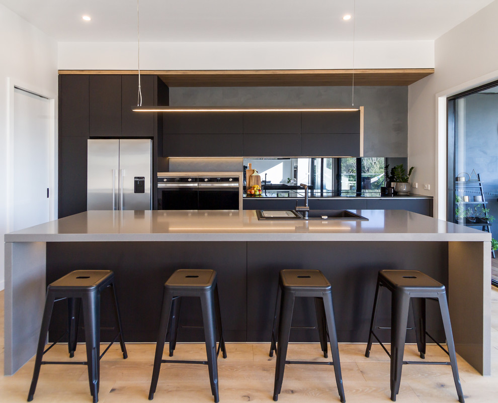 Design ideas for a mid-sized industrial galley kitchen in Melbourne with a drop-in sink, flat-panel cabinets, black cabinets, quartz benchtops, black splashback, mirror splashback, stainless steel appliances, light hardwood floors, beige floor, grey benchtop and a peninsula.
