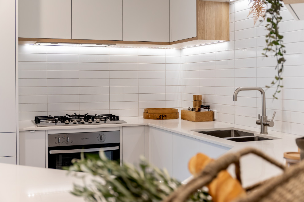 Small contemporary u-shaped eat-in kitchen in Adelaide with an undermount sink, flat-panel cabinets, white cabinets, quartz benchtops, white splashback, subway tile splashback, stainless steel appliances, laminate floors, a peninsula, brown floor and white benchtop.