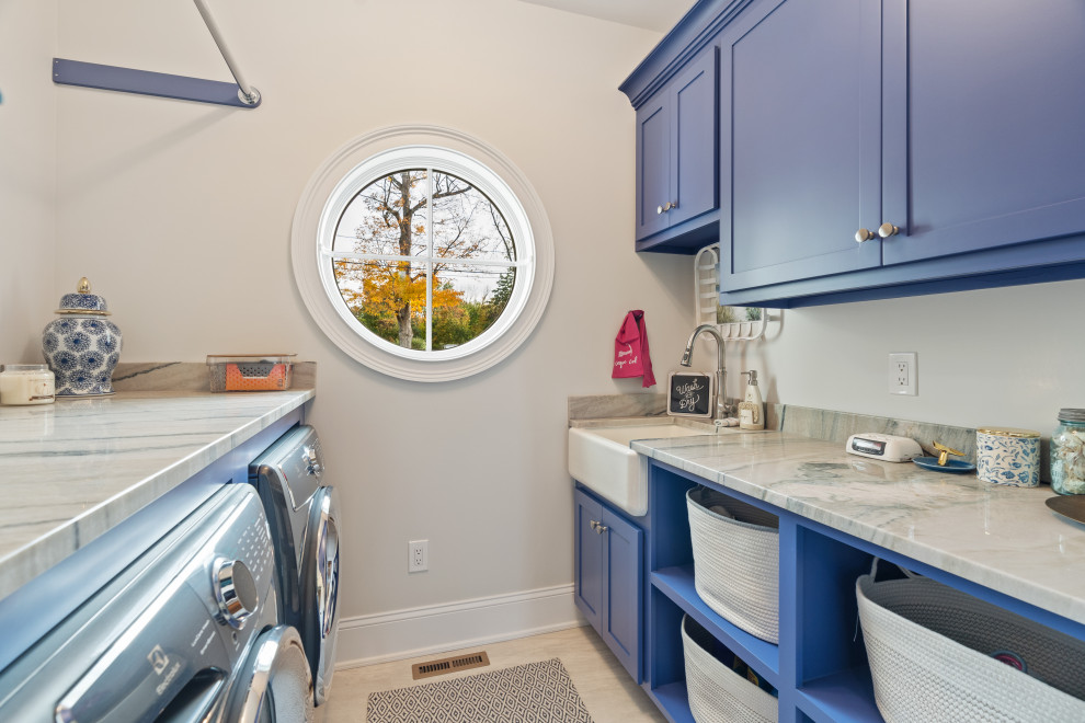 This is an example of a large country galley dedicated laundry room in Bridgeport with blue cabinets, marble benchtops, grey splashback, grey walls, light hardwood floors, a side-by-side washer and dryer, beige floor and white benchtop.