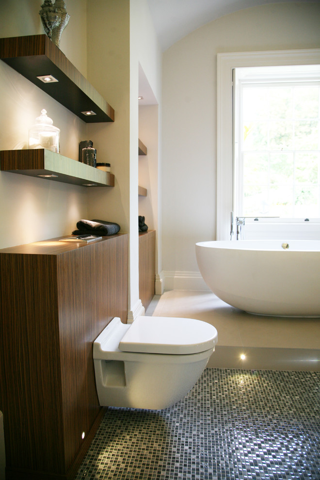 Photo of a large contemporary master bathroom in Other with flat-panel cabinets, medium wood cabinets, a freestanding tub, a wall-mount toilet, multi-coloured tile, mosaic tile, beige walls, mosaic tile floors, a vessel sink and glass benchtops.