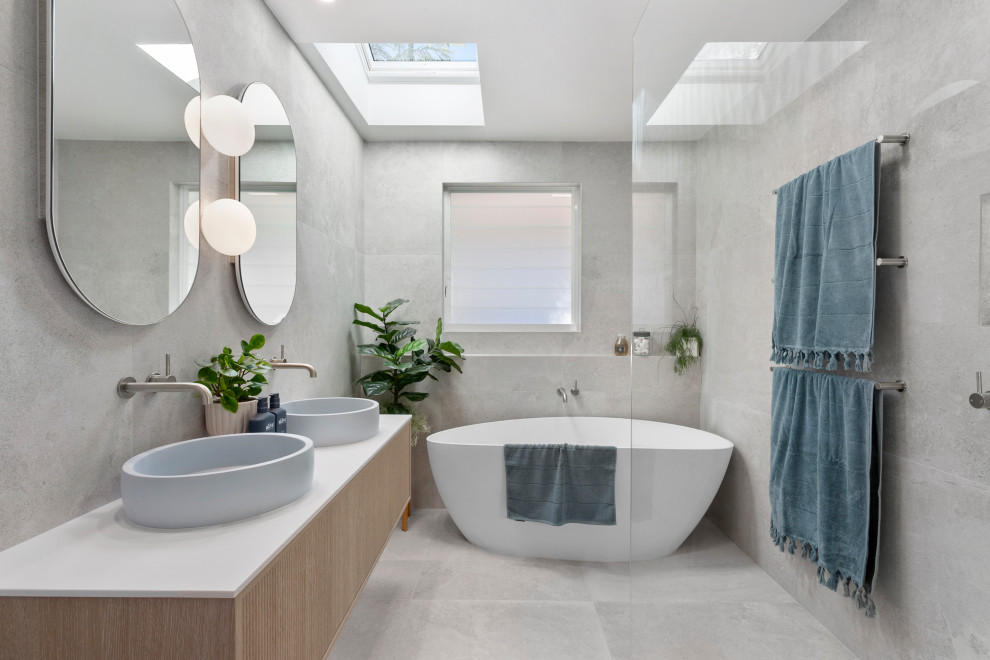 Idéer för stora funkis vitt en-suite badrum, med skåp i ljust trä, ett fristående badkar, en öppen dusch, en toalettstol med hel cisternkåpa, grå kakel, keramikplattor, grå väggar, klinkergolv i keramik, ett fristående handfat, bänkskiva i akrylsten, grått golv, med dusch som är öppen och släta luckor