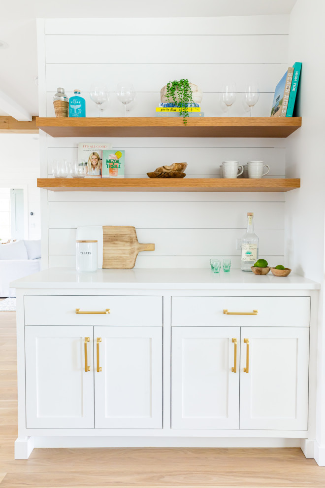 This is an example of a large beach style l-shaped eat-in kitchen in Other with a farmhouse sink, shaker cabinets, white cabinets, quartz benchtops, white splashback, shiplap splashback, panelled appliances, light hardwood floors, with island, brown floor and white benchtop.