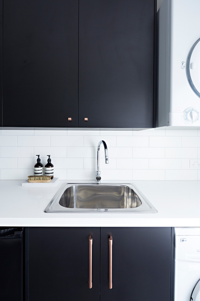 Mid-sized modern single-wall dedicated laundry room in Melbourne with an undermount sink, black cabinets, a stacked washer and dryer, black floor, white benchtop, quartz benchtops and white walls.