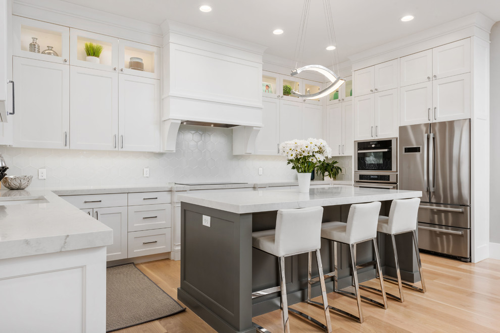This is an example of a transitional u-shaped kitchen in Salt Lake City with shaker cabinets, white cabinets, white splashback, stainless steel appliances, light hardwood floors, with island and grey benchtop.