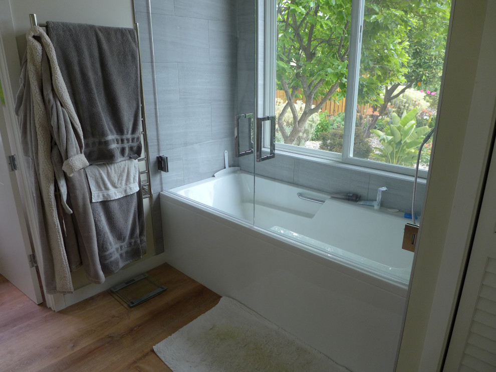 Design ideas for a mid-sized tropical master bathroom in San Francisco with flat-panel cabinets, white cabinets, an alcove tub, a shower/bathtub combo, a one-piece toilet, white tile, porcelain tile, grey walls, light hardwood floors, a drop-in sink, engineered quartz benchtops, brown floor, a hinged shower door, white benchtops, an enclosed toilet, a double vanity and a built-in vanity.
