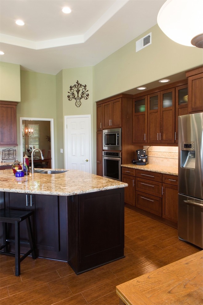 Transitional l-shaped porcelain tile kitchen photo with an undermount sink, recessed-panel cabinets, medium tone wood cabinets, granite countertops, beige backsplash, stone tile backsplash, stainless steel appliances and an island