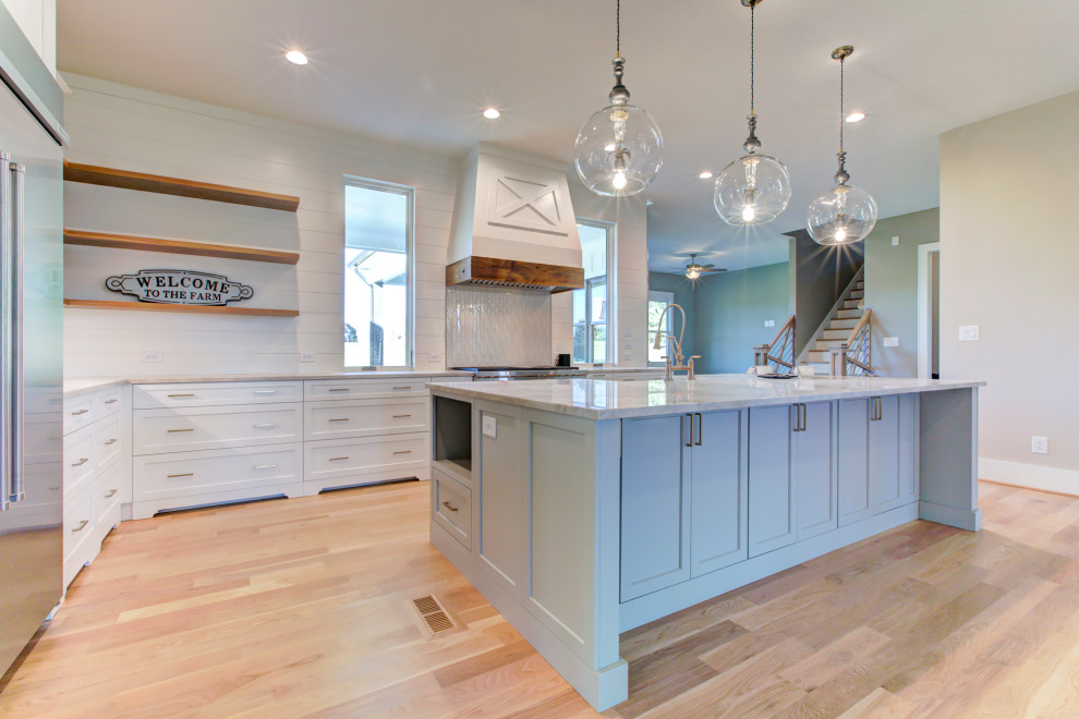 Design ideas for a large farmhouse l-shaped kitchen in Raleigh with a belfast sink, recessed-panel cabinets, quartz worktops, metallic splashback, glass tiled splashback, stainless steel appliances, light hardwood flooring, an island and white worktops.