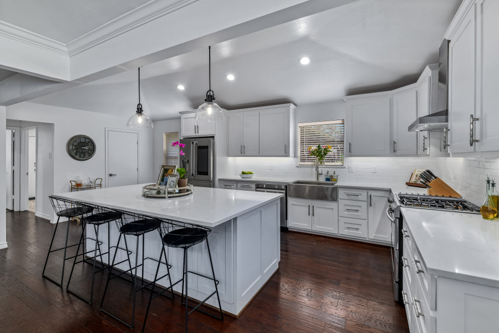 Large contemporary l-shaped eat-in kitchen in Dallas with a farmhouse sink, shaker cabinets, white cabinets, quartz benchtops, white splashback, ceramic splashback, stainless steel appliances, medium hardwood floors, with island, brown floor, white benchtop and vaulted.