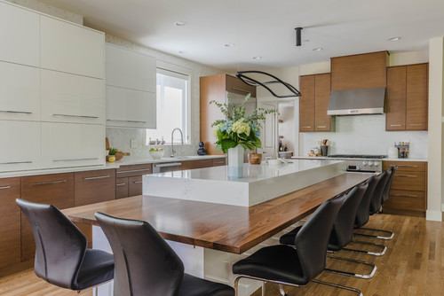Modern Canadian kitchen with two-tone slab cabinets (white uppers and warm wood lowers), a long island with waterfall quartz and wood dining ledge, stainless range hood, and black bar stools