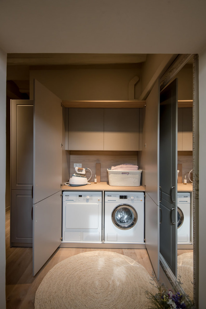 This is an example of a small transitional single-wall laundry cupboard in Barcelona with flat-panel cabinets, a side-by-side washer and dryer, wood benchtops, light hardwood floors, beige floor, beige benchtop and grey cabinets.