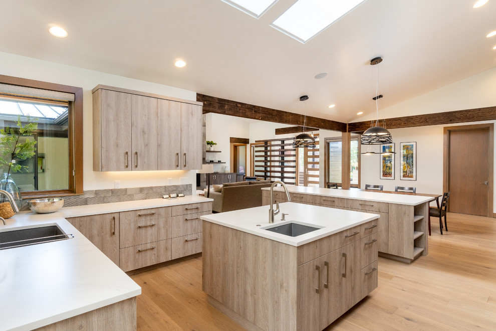 Large mountain style u-shaped light wood floor open concept kitchen photo with an undermount sink, flat-panel cabinets, light wood cabinets, quartz countertops, gray backsplash, ceramic backsplash, paneled appliances, two islands and white countertops