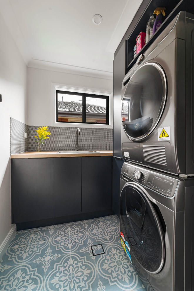 Foto di una lavanderia minimal con lavello da incasso, ante lisce, ante nere, top in legno, pareti bianche, lavatrice e asciugatrice a colonna, pavimento blu e top marrone
