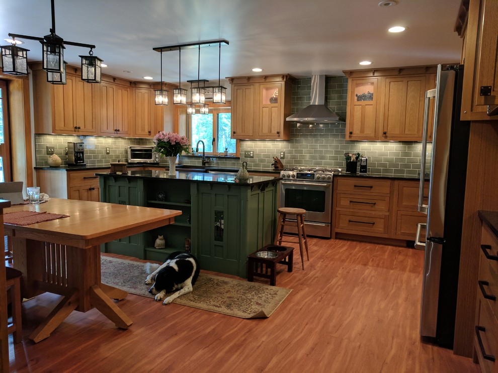 Photo of a large arts and crafts u-shaped eat-in kitchen in New York with a farmhouse sink, shaker cabinets, medium wood cabinets, solid surface benchtops, green splashback, subway tile splashback, stainless steel appliances, medium hardwood floors, with island, brown floor and black benchtop.