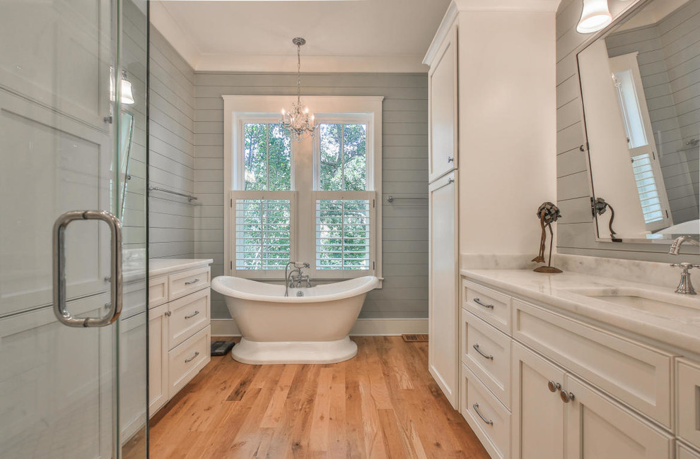 Photo of a beach style 3/4 bathroom in Charleston with recessed-panel cabinets, white cabinets, a freestanding tub, grey walls, medium hardwood floors, an undermount sink and brown floor.
