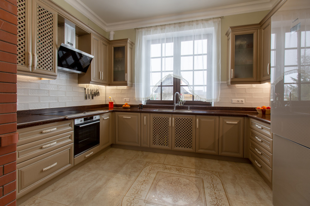 Photo of a large traditional u-shaped eat-in kitchen in Saint Petersburg with a double-bowl sink, raised-panel cabinets, brown cabinets, solid surface benchtops, white splashback, subway tile splashback, black appliances, ceramic floors, no island, beige floor and brown benchtop.