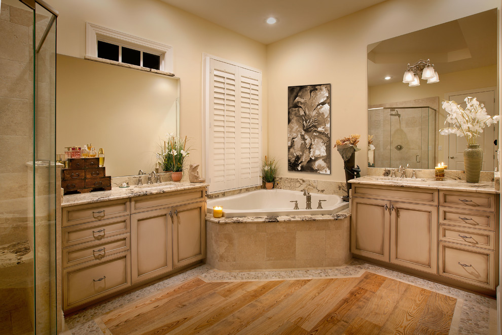 Inspiration for a large traditional master bathroom in Miami with recessed-panel cabinets, beige cabinets, a corner tub, a corner shower, a two-piece toilet, beige tile, limestone, beige walls, medium hardwood floors, an undermount sink, granite benchtops, brown floor and a hinged shower door.