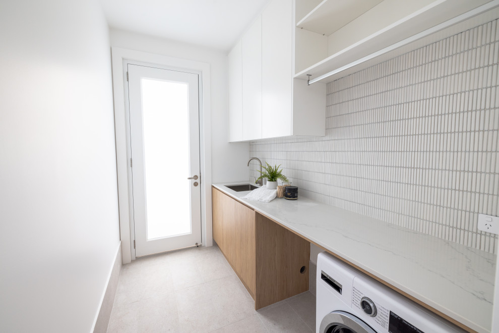 This is an example of a beach style laundry room in Central Coast with an undermount sink, flat-panel cabinets, light wood cabinets, quartz benchtops, white splashback, mosaic tile splashback, white walls, porcelain floors, a side-by-side washer and dryer, grey floor and white benchtop.