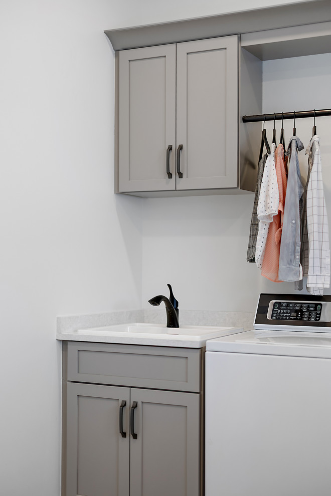 This is an example of a large transitional l-shaped dedicated laundry room in Detroit with a drop-in sink, shaker cabinets, grey cabinets, quartz benchtops, white splashback, engineered quartz splashback, white walls, porcelain floors, a side-by-side washer and dryer, grey floor and white benchtop.