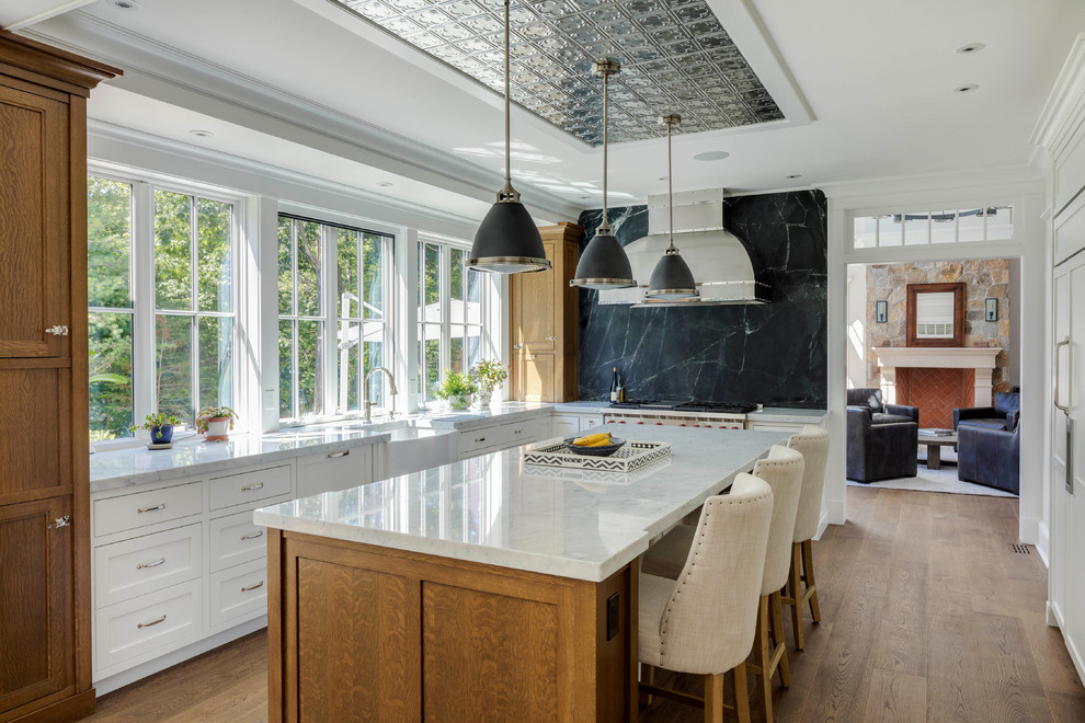 Example of a large classic l-shaped medium tone wood floor and brown floor enclosed kitchen design in Boston with a farmhouse sink, shaker cabinets, white cabinets, marble countertops, black backsplash, stone slab backsplash, stainless steel appliances, an island and gray countertops