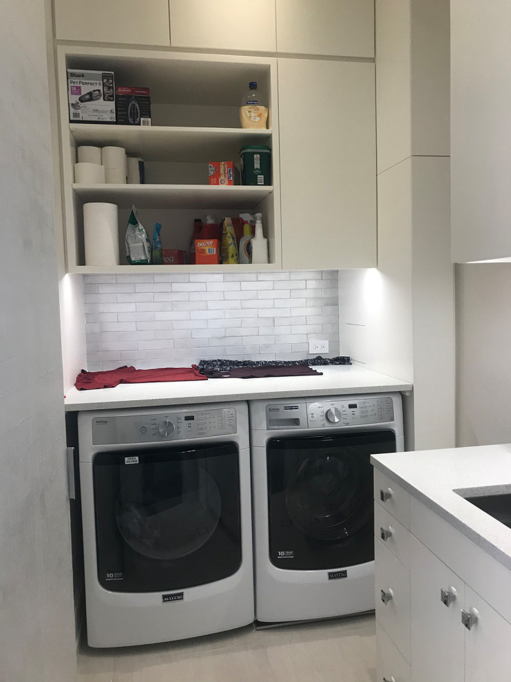 Photo of a mid-sized transitional l-shaped dedicated laundry room in New York with an undermount sink, flat-panel cabinets, white cabinets, quartz benchtops, white splashback, porcelain splashback, grey walls, porcelain floors, a side-by-side washer and dryer, grey floor and white benchtop.