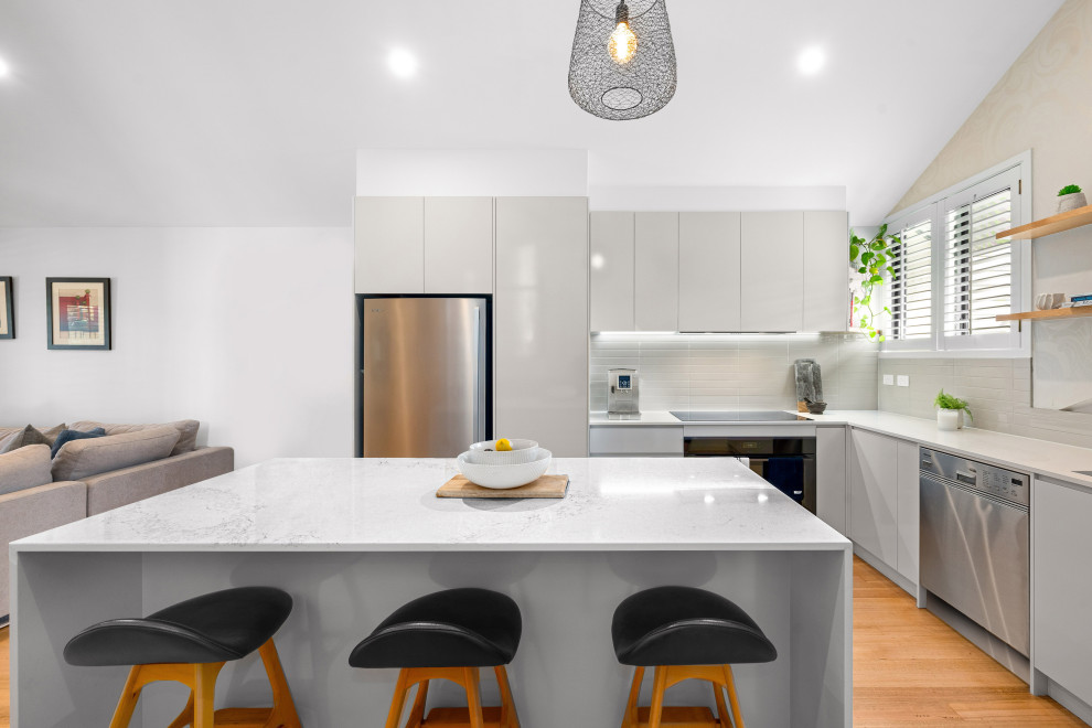 Design ideas for a contemporary l-shaped kitchen in Brisbane with flat-panel cabinets, grey cabinets, grey splashback, stainless steel appliances, medium hardwood floors, with island, brown floor and white benchtop.