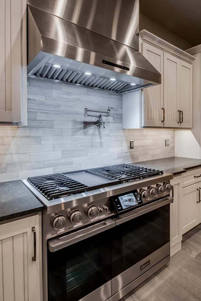 Photo of a large traditional single-wall kitchen in Denver with a submerged sink, shaker cabinets, distressed cabinets, granite worktops, grey splashback, limestone splashback, stainless steel appliances, light hardwood flooring, multiple islands, grey floors and black worktops.