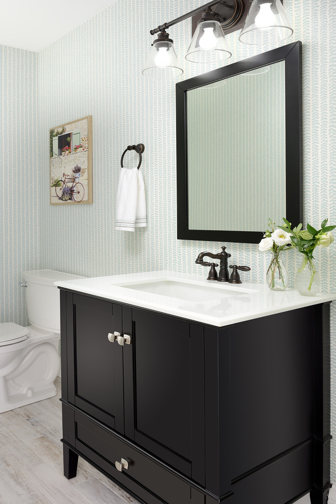 Inspiration for a mid-sized beach style powder room in Toronto with recessed-panel cabinets, brown cabinets, a two-piece toilet, multi-coloured walls, laminate floors, a drop-in sink, engineered quartz benchtops, grey floor and white benchtops.