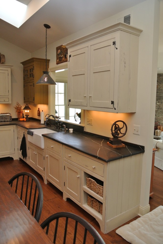 Large country l-shaped open plan kitchen in New York with a farmhouse sink, recessed-panel cabinets, beige cabinets, soapstone benchtops, black splashback, stainless steel appliances, medium hardwood floors and with island.