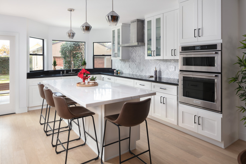 Large transitional u-shaped eat-in kitchen in Los Angeles with an undermount sink, shaker cabinets, white cabinets, marble benchtops, grey splashback, marble splashback, stainless steel appliances, light hardwood floors, with island, brown floor and white benchtop.