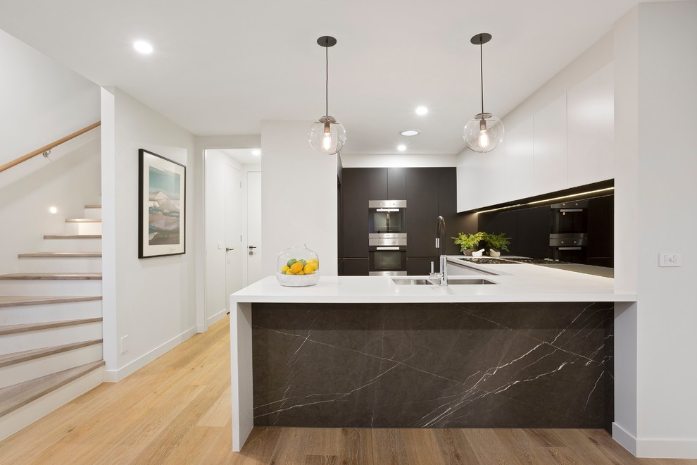 Inspiration for a contemporary u-shaped kitchen in Melbourne with a double-bowl sink, flat-panel cabinets, grey cabinets, black splashback, glass sheet splashback, stainless steel appliances, medium hardwood floors, a peninsula, brown floor and white benchtop.