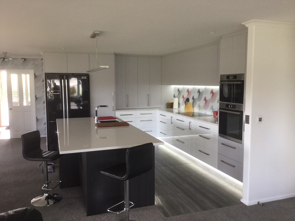 This is an example of a large contemporary l-shaped open plan kitchen in Christchurch with a single-bowl sink, flat-panel cabinets, white cabinets, quartz benchtops, multi-coloured splashback, glass sheet splashback, stainless steel appliances, vinyl floors and with island.