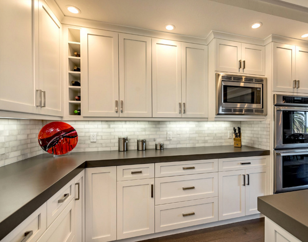 Example of a mid-sized transitional l-shaped medium tone wood floor enclosed kitchen design in Detroit with an island, shaker cabinets, white cabinets, quartz countertops, gray backsplash, stone tile backsplash and stainless steel appliances