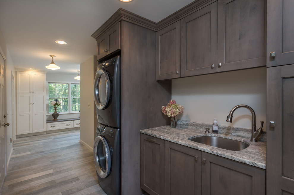 Inspiration for a mid-sized country single-wall utility room in Boston with an undermount sink, recessed-panel cabinets, grey cabinets, granite benchtops, porcelain floors, a stacked washer and dryer and grey floor.
