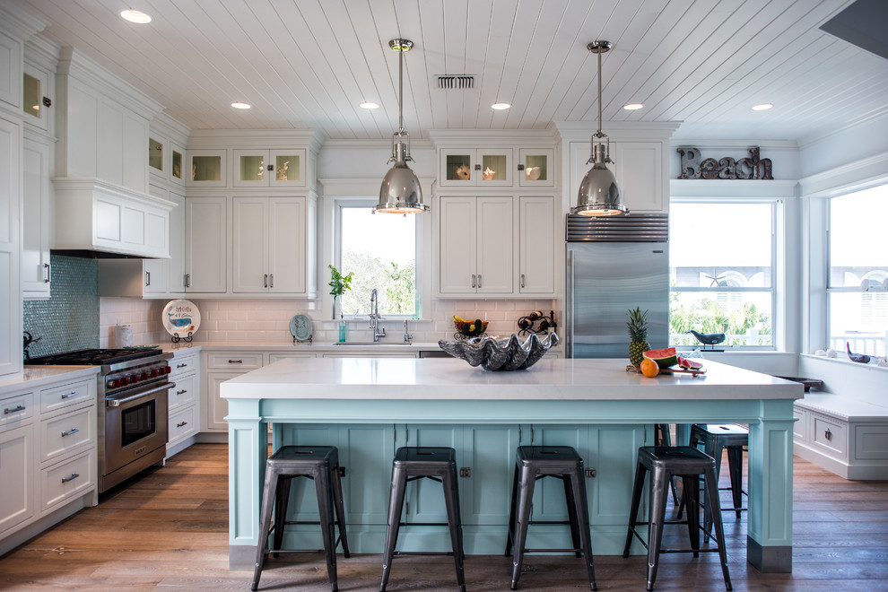 Design ideas for a mid-sized beach style l-shaped open plan kitchen in Other with shaker cabinets, white cabinets, white splashback, subway tile splashback, stainless steel appliances, with island, an undermount sink, solid surface benchtops, medium hardwood floors and brown floor.