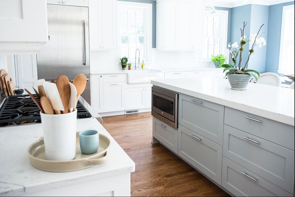 Example of a large transitional l-shaped medium tone wood floor and brown floor open concept kitchen design in Chicago with a farmhouse sink, recessed-panel cabinets, white cabinets, quartz countertops, white backsplash, subway tile backsplash, stainless steel appliances, an island and white countertops