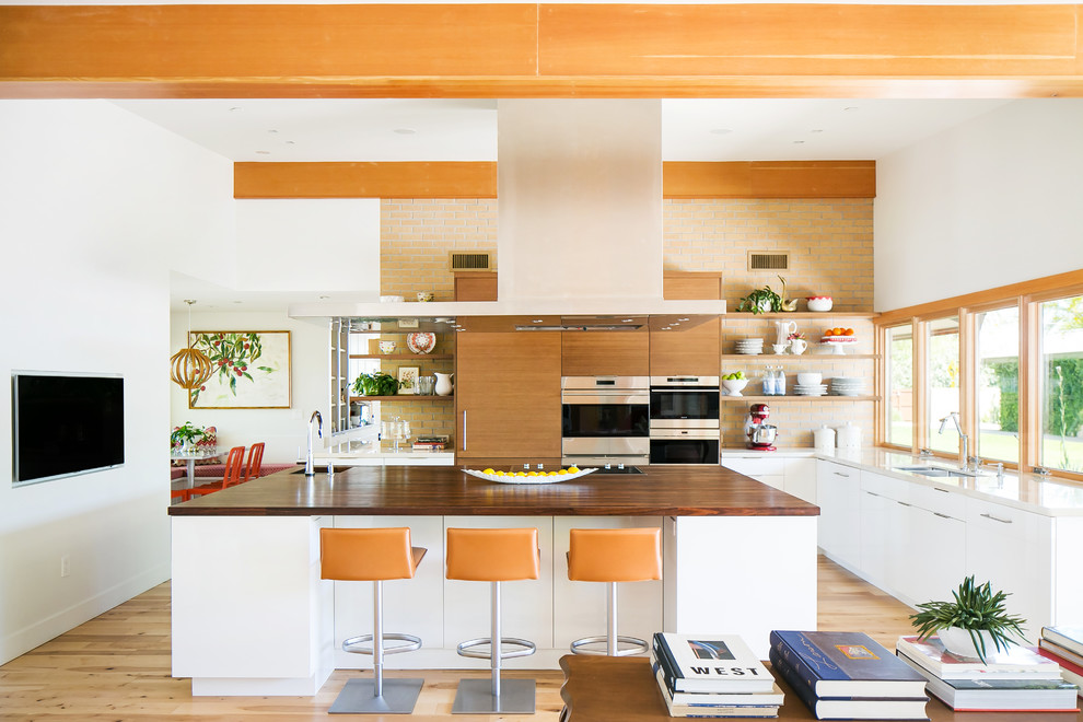 Design ideas for a midcentury l-shaped open plan kitchen in Phoenix with light hardwood floors, with island, a double-bowl sink, flat-panel cabinets, medium wood cabinets, wood benchtops, orange splashback, brick splashback, stainless steel appliances, beige floor and brown benchtop.