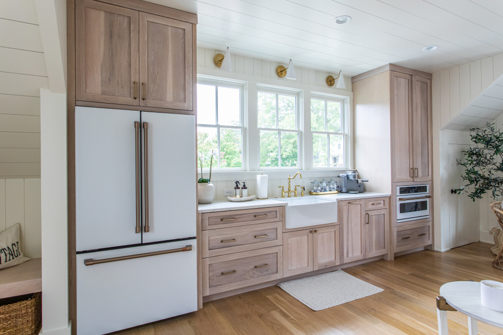 Single-wall kitchen in Atlanta with a farmhouse sink, shaker cabinets, light wood cabinets, timber splashback, white appliances, light hardwood floors, brown floor, white benchtop and timber.