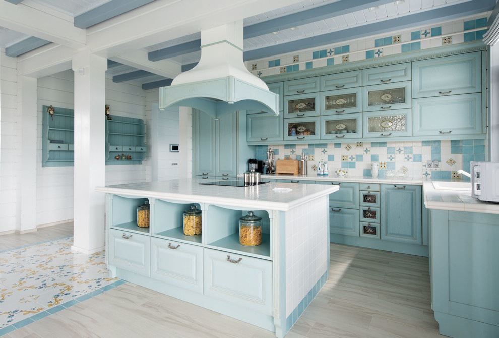 Mediterranean l-shaped open plan kitchen with a drop-in sink, raised-panel cabinets, turquoise cabinets, tile benchtops, multi-coloured splashback, ceramic splashback, with island, beige floor and white benchtop.