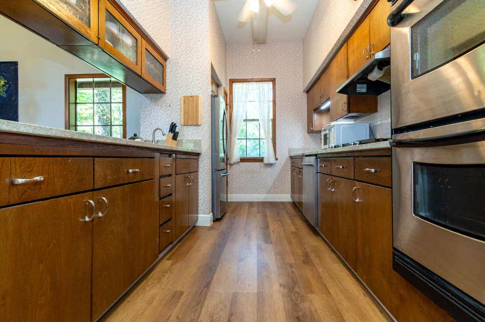 This is an example of a mid-sized country galley separate kitchen in New Orleans with a single-bowl sink, flat-panel cabinets, medium wood cabinets, marble benchtops, grey splashback, cement tile splashback, stainless steel appliances, vinyl floors, brown floor, beige benchtop and wallpaper.
