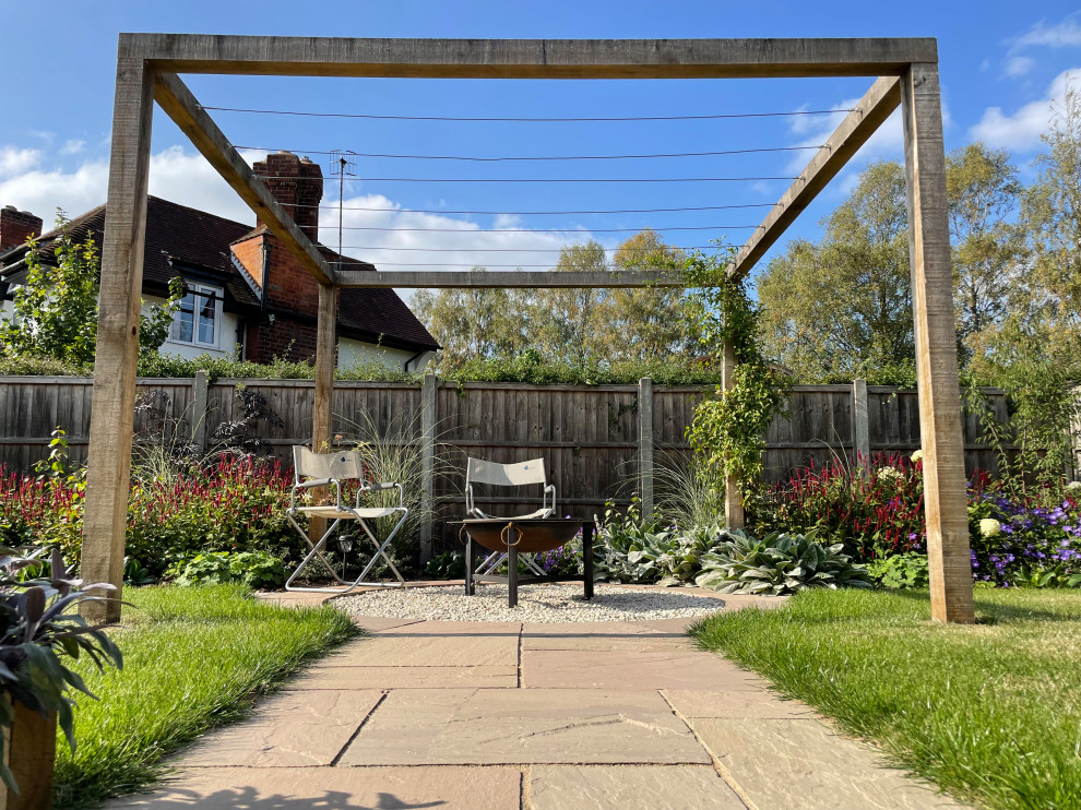 Aménagement d'un jardin avec pergola arrière campagne l'été avec une exposition ensoleillée, des pavés en pierre naturelle et une clôture en bois.