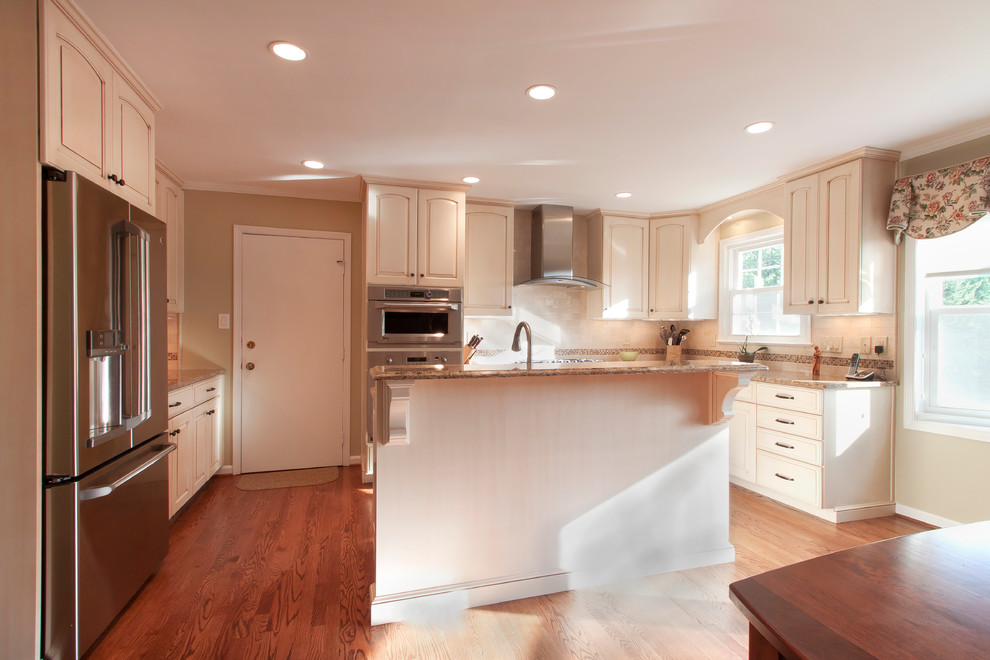 This is an example of a traditional u-shaped eat-in kitchen in DC Metro with raised-panel cabinets, quartz benchtops, beige splashback, stainless steel appliances, medium hardwood floors and with island.