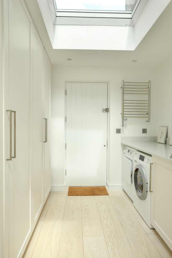 Inspiration for a large modern galley dedicated laundry room in London with shaker cabinets, solid surface benchtops, white walls, light hardwood floors and white benchtop.