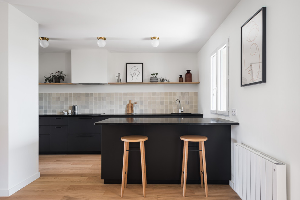 Inspiration for a mid-sized contemporary single-wall open plan kitchen in Bordeaux with black cabinets, granite benchtops, grey splashback, ceramic splashback, with island, black benchtop, an undermount sink, flat-panel cabinets, medium hardwood floors and brown floor.