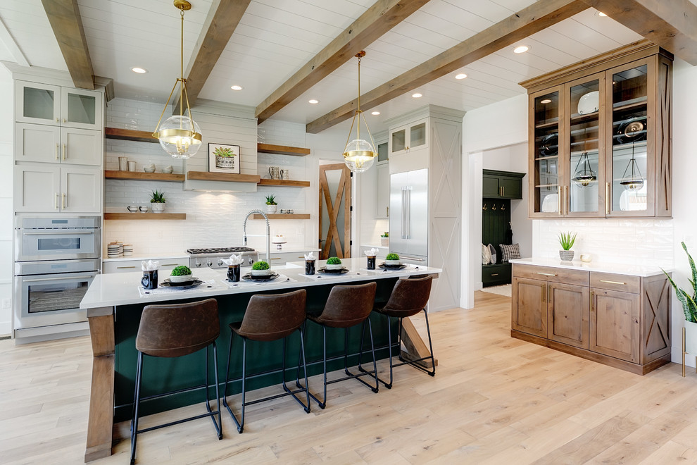 This is an example of a large country l-shaped open plan kitchen in Boise with a farmhouse sink, shaker cabinets, white cabinets, quartz benchtops, white splashback, subway tile splashback, stainless steel appliances, light hardwood floors, with island, beige floor and white benchtop.