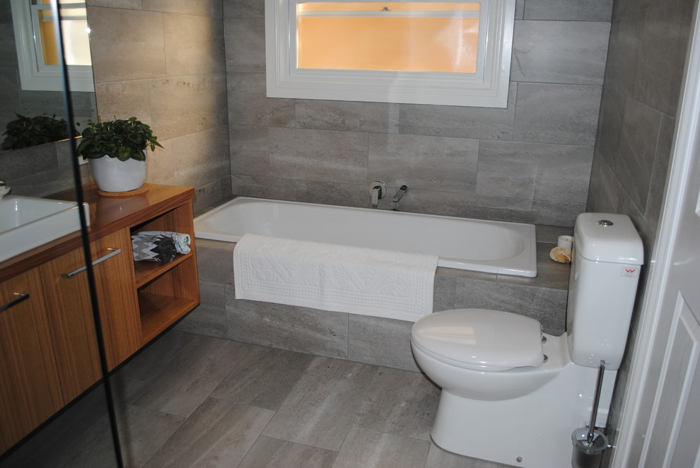 This is an example of a mid-sized contemporary master bathroom in Other with furniture-like cabinets, light wood cabinets, a drop-in tub, an open shower, a two-piece toilet, gray tile, ceramic tile, grey walls, ceramic floors, a drop-in sink, wood benchtops, grey floor and an open shower.