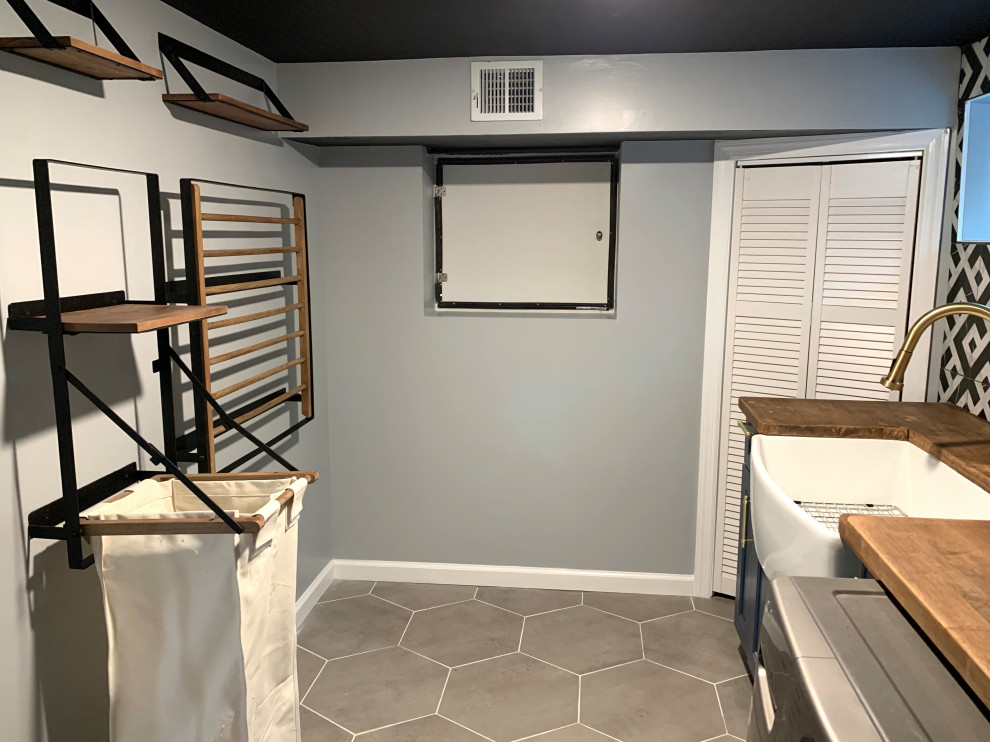 This is an example of a mid-sized contemporary galley dedicated laundry room in New York with a farmhouse sink, beaded inset cabinets, blue cabinets, wood benchtops, multi-coloured splashback, cement tile splashback, grey walls, ceramic floors, a side-by-side washer and dryer, grey floor and brown benchtop.