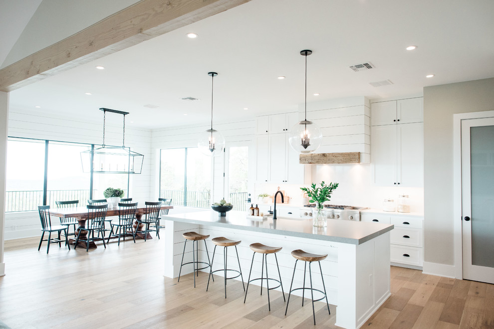 Photo of a large transitional u-shaped open plan kitchen in Austin with a farmhouse sink, shaker cabinets, white cabinets, quartzite benchtops, white splashback, stone slab splashback, stainless steel appliances, light hardwood floors, with island, brown floor and grey benchtop.