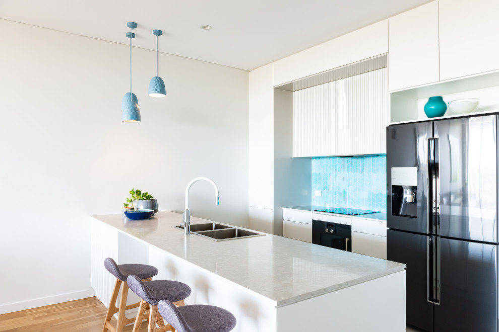 This is an example of a contemporary galley kitchen in Adelaide with a double-bowl sink, flat-panel cabinets, white cabinets, blue splashback, mosaic tile splashback, stainless steel appliances, medium hardwood floors, with island, brown floor and white benchtop.