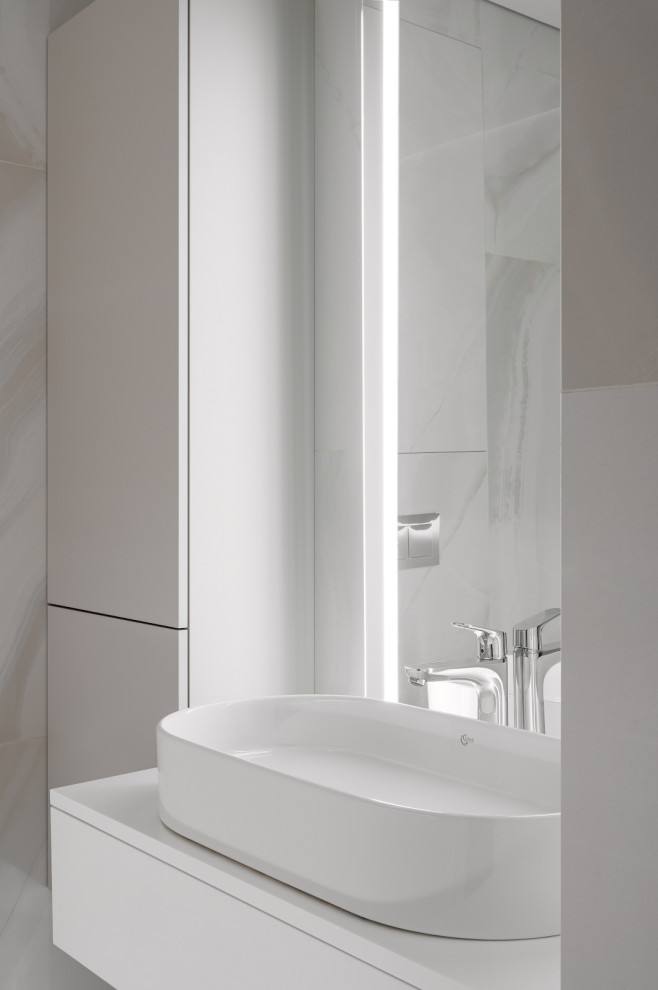 Пример оригинального дизайна: главная, серо-белая ванная комната среднего размера в современном стиле с плоскими фасадами, инсталляцией, полом из керамогранита, настольной раковиной, столешницей из искусственного камня, белой столешницей, тумбой под одну раковину, подвесной тумбой, белыми фасадами, полновстраиваемой ванной, серой плиткой, керамогранитной плиткой, серыми стенами, серым полом и зеркалом с подсветкой